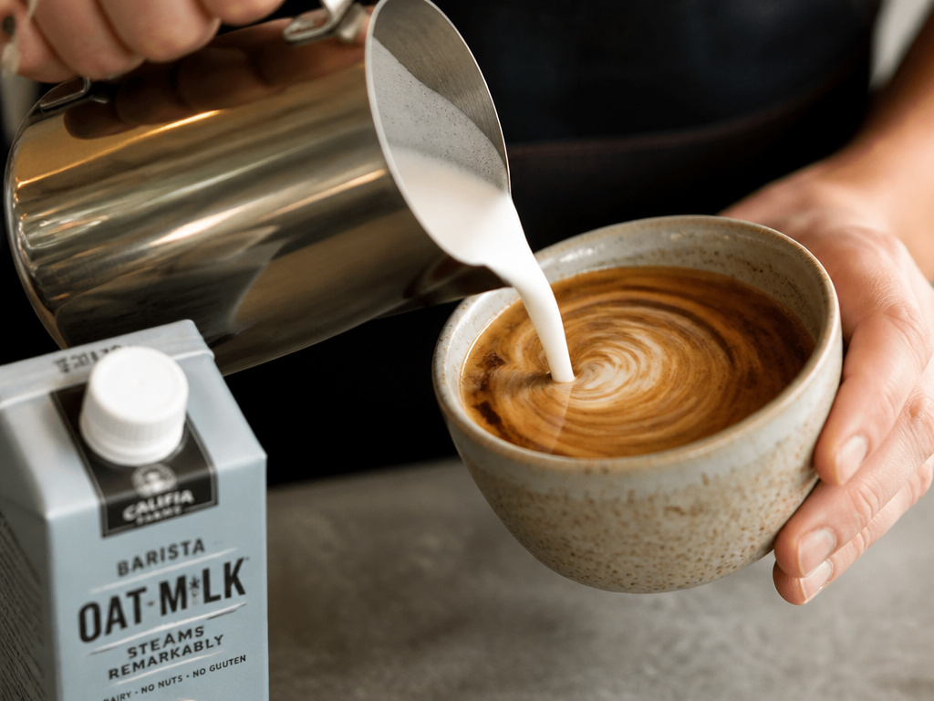 Steamed milk being poured into espresso for a latte, with dairy and oat milk cartons visible.