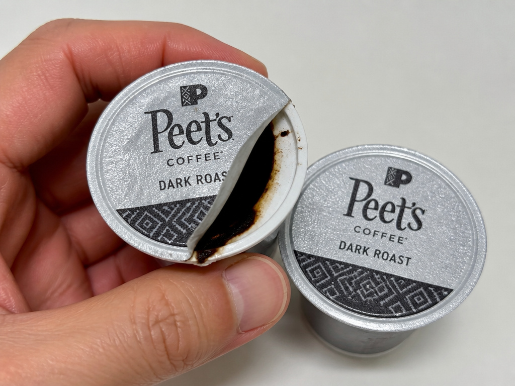 Close-up of a damaged coffee pod with a torn foil lid and oil stain.